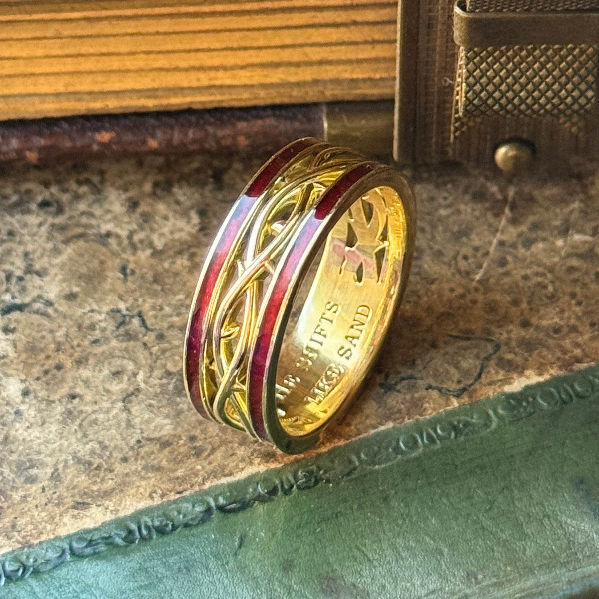 A gold band ring with intricate Celtic knotwork engravings and red enamel inlay, resting on a textured vintage book page.