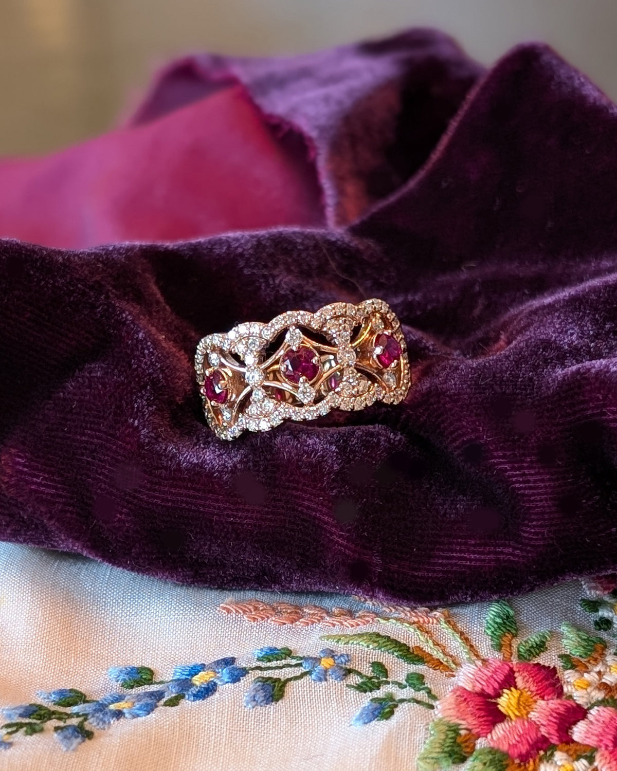 An ornate rose gold ring with three red Rubies and pavé diamond accents, displayed on draped deep purple velvet fabric.