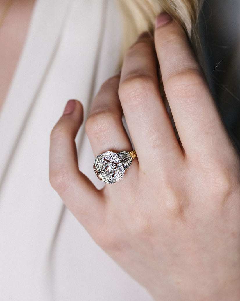 A close-up of a hand wearing a vintage-style diamond ring with a gold band, set against a soft cream-colored dress.
