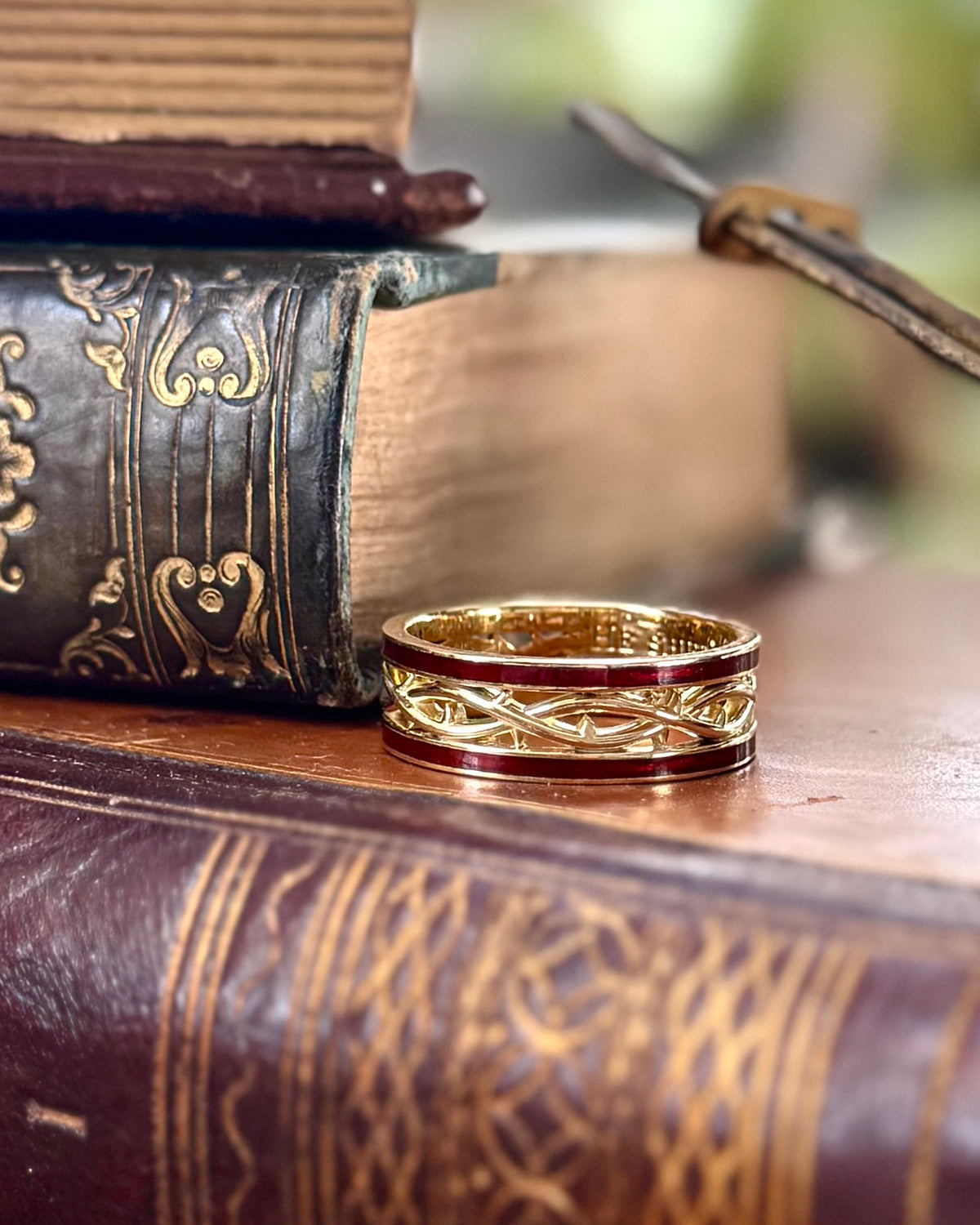 A gold ring with a red enamel band and intricate Celtic knotwork detailing, resting on a wooden surface next to antique books.