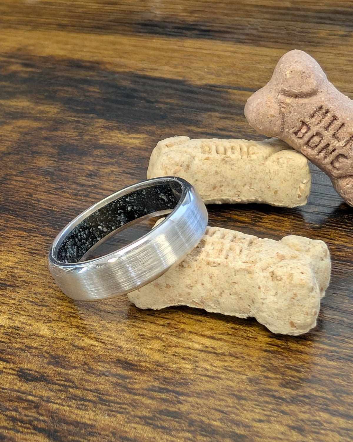 A brushed white gold wedding band with a black enameled interior that contains the client's dog's ashes.  The ring is displayed on a dark wood surface next to dog treats.