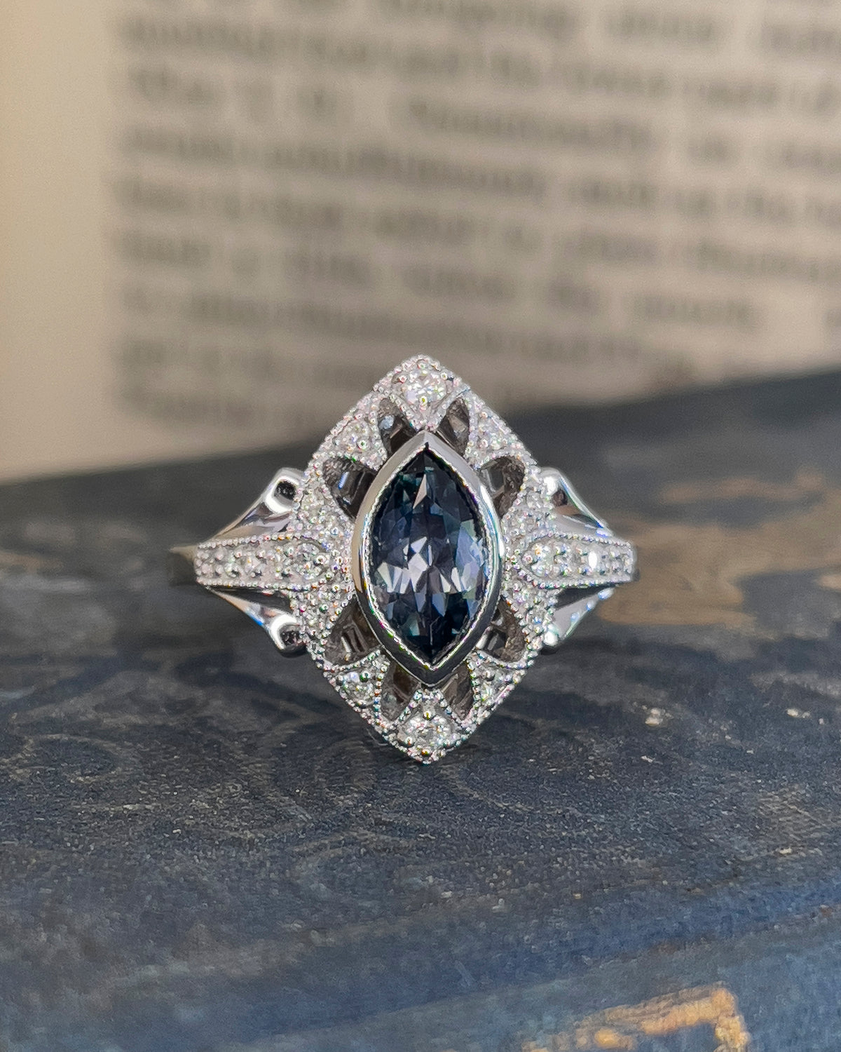 An ornate white gold ring featuring a marquise-cut Montana Blue Sapphire surrounded by small round diamonds, displayed on a dark blue book.