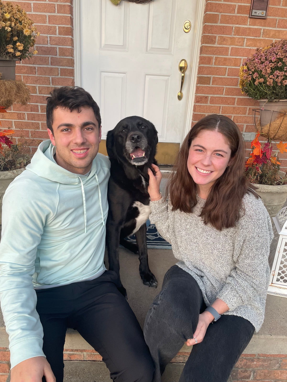 A man and a woman sitting together on the front steps of a brick house, with a black dog between them. The man wears a light blue hoodie and black pants, and the woman wears a light gray knit sweater and dark jeans.