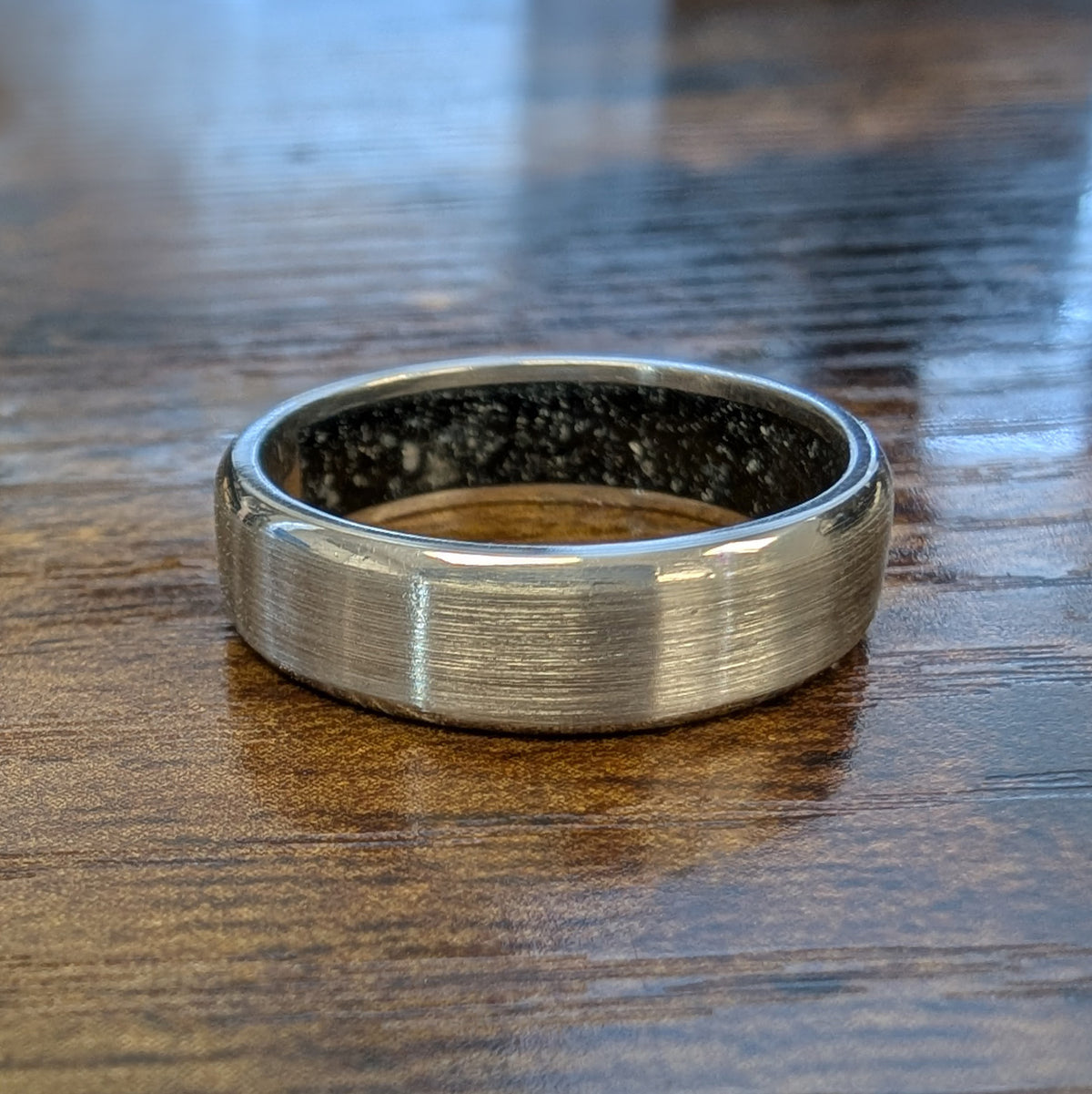 A brushed white gold gents' wedding ring with a black enamel interior containing the dog's ashes, resting on a dark wood tabletop.