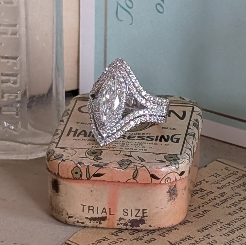 A white gold halo ring with a marquise-cut center stone and surrounding round stones, displayed on top of a vintage floral hair dressing box.