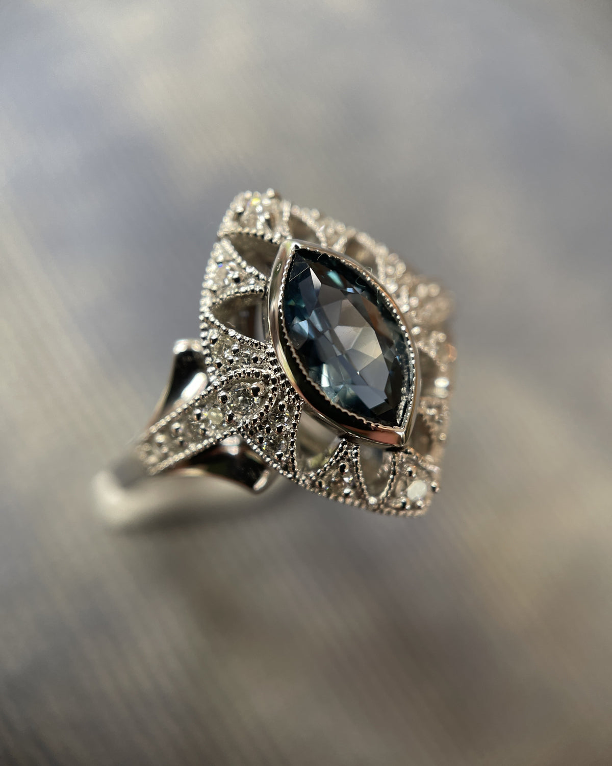 An ornate white gold ring featuring a marquise-cut dark Montane Blue Sapphire surrounded by pavé-set diamonds, photographed on a blurred gray background.